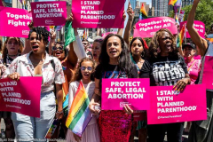 A woman with a t-shirt saying "1973" and a sign saying "Protect safe, legal abortion - Planned Parenthood" and other people holding signs saying "I stand with Planned Parenthood" marching with the Planned Parenthood group at the New York City Pride March.
